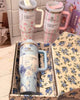 Two floral-patterned tumblers with pink handles on a floral-patterned surface.