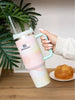 Hand holding a colorful Stanley tumbler with a plant and pastry in the background